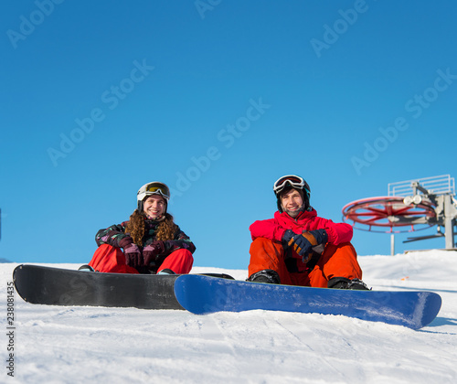 Guy and girl snowboarders are sitting on the snow at the top of the ski slope against the blue sky on a sunny day. Couple looking at camera and smiling © anatoliy_gleb