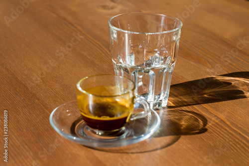 A transparent cup of coffee ground and an empty glass stand on the table. The morning sun shines on the cup and glass, their shadows on the table.