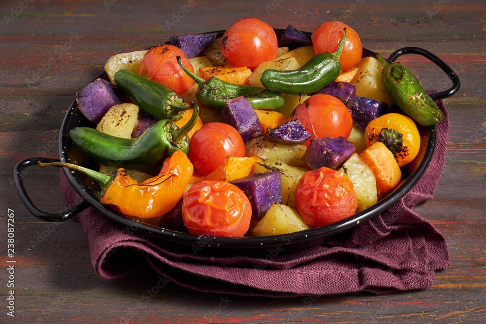 Rustic, oven baked vegetables in baking dish. Seasonal vegetarian vegan meal on dark wooden background with linen towel