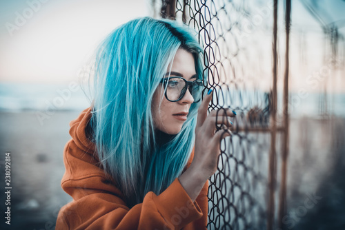 Portrait of young hipster woman with blue hair and glasses sitting on beach near mesh fence photo