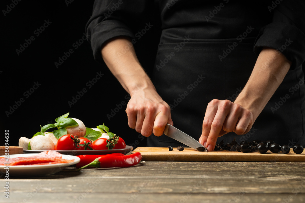 The chef cuts the olives. For the preparation of pizza, salad. A delicious meal concept. On a black background for design or lettering text. The chef cuts the olives. For the preparation of pizza, salad. A delicious meal concept. On a black background for design or lettering text.