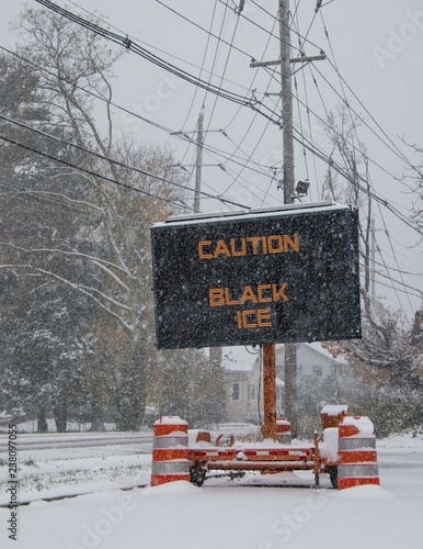 Electric road traffic mobile sign by the side of a snow covered road with snow falling warning of black ice on road