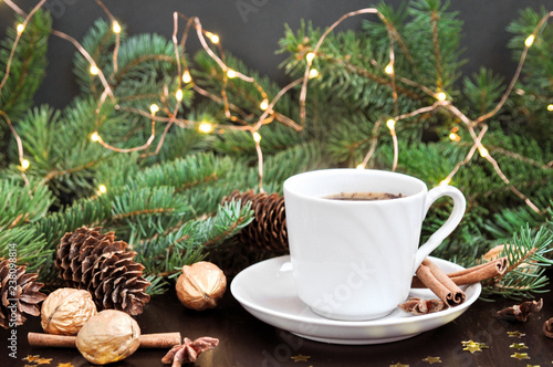 Winter season holidays mood. Cup of coffee with cinnamon surrounded by fir branches and cones with lights garland on rustic wooden background. flat lay