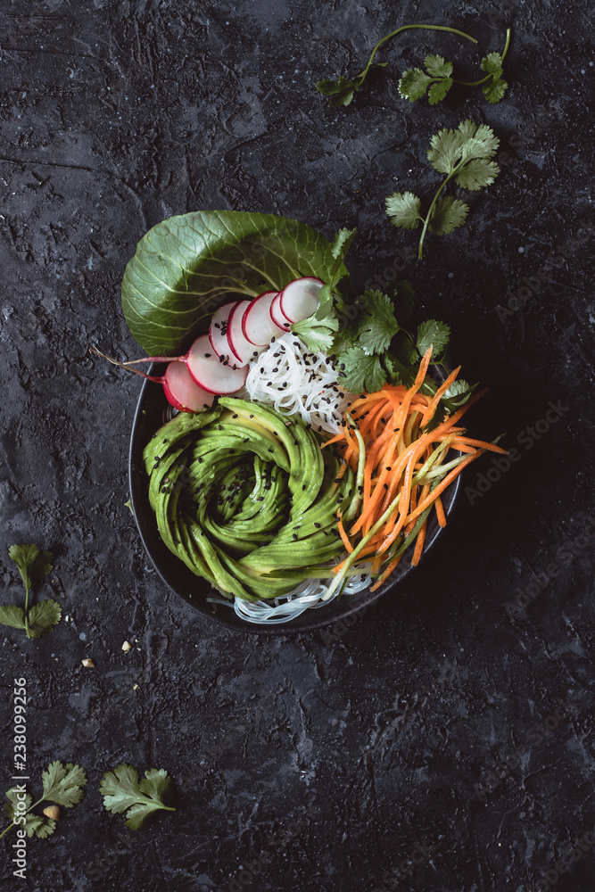 Obraz premium Raw vegan bowl with rice noodles, carrot, radish and avocado on black background. Top view