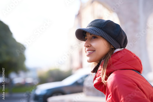 Portrait of a young smiling woman in red jacket,gray scarf and cap. Woman posing outdoors on the street at sunny autumn or spring day © vitaliymateha