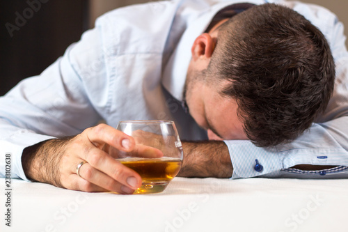 Pijany mężczyzna śpi przy stole z głową opartą o stół. W ręce trzyma szklankę whisky.