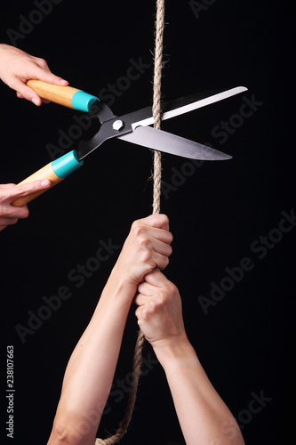A person hanging from a rope and another one cutting it with big scissors.