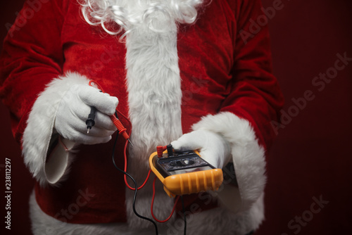 Closeup on Santa Claus solving electricity problems using holding digital tester multimeter electronic measure equipment. Ready for Merry Christmas and Happy New Year celebration background.