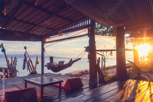 Obraz na płótnie Young attractive guy a man with a beard in a hammock with a laptop on the background of the sea and sunset, distant work, freelancer, blogger, vacation and travel w salonie