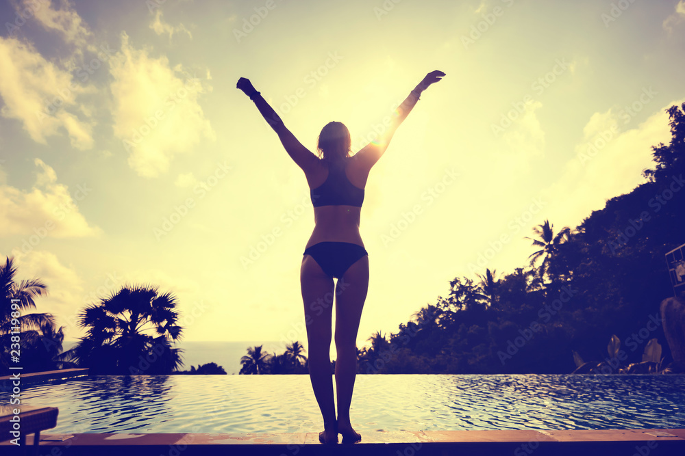 Fototapeta premium Silhouette of a woman in a bikini on the side of an infinity pool overlooking the sunset jungle and tropical sea, luxury holidays and vacations in hot countries
