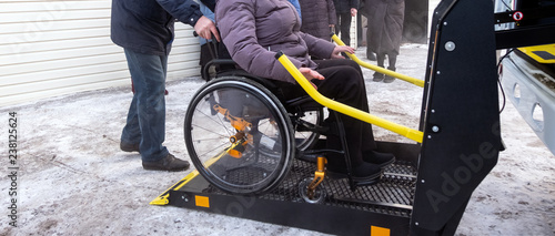 A woman in a wheelchair on a lift of a specialized vehicle for people with disabilities. Taxi for the disabled. Yellow bar and handrail. Winter.