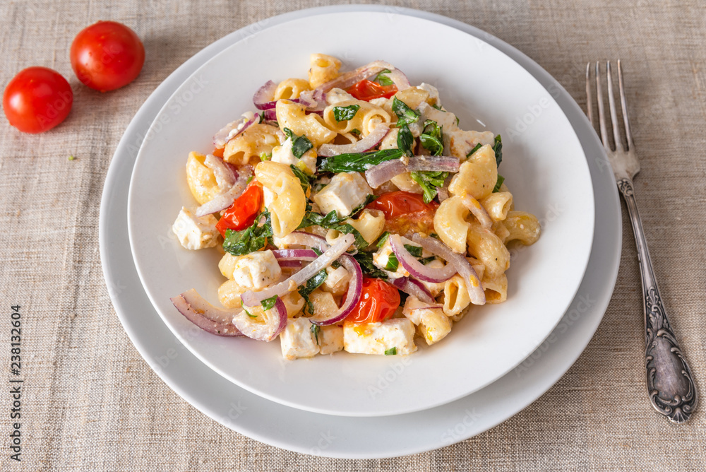 Warm pasta salad, cherry tomatoes, feta cheese and onions on a plate, top view, rustic background Warm pasta salad, cherry tomatoes, feta cheese and onions on a plate, top view, rustic background