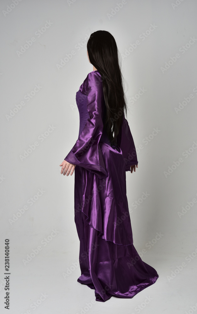 full length portrait of beautiful girl with long black hair,   wearing purple fantasy medieval gown. standing pose with back to the camera on grey studio background.