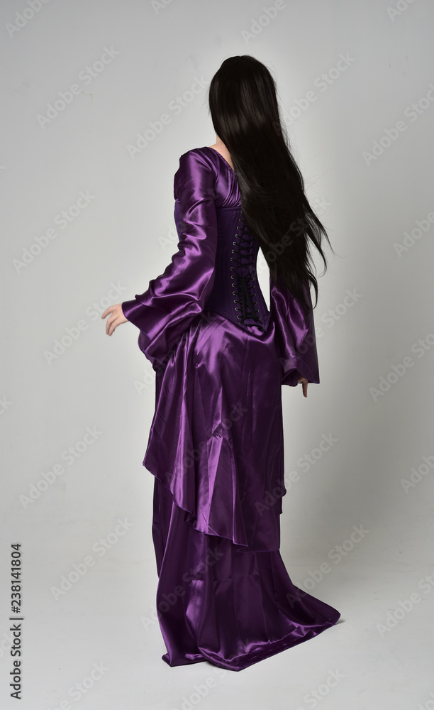 full length portrait of beautiful girl with long black hair, wearing purple fantasy medieval gown. standing pose with back to the camera on grey studio background. full length portrait of beautiful girl with long black hair, wearing purple fantasy medieval gown. standing pose with back to the camera on grey studio background.