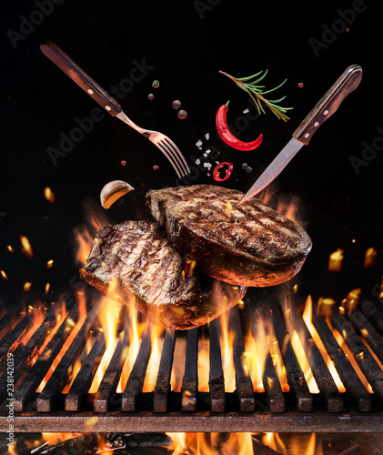 Raw beef steaks with vegetables and spices fly over the blazing grill barbecue fire.
