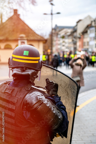 Rear view of police officer wearing helmet and shield securing the zone in frong of the Yellow vests movement gilets jaunes protesters on Quai des Bateliers street . photo