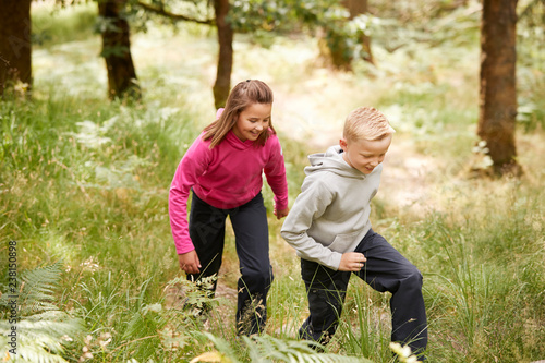 Two children walking together in a forest amongst greenery, three quarter length, side view