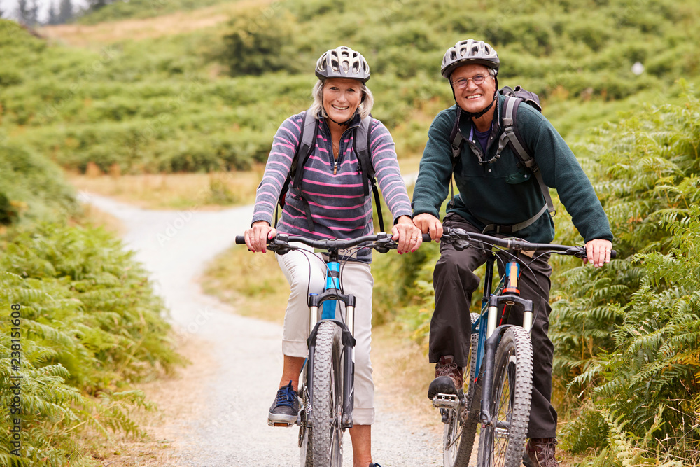 Senior couple sitting on mountain bikes in a country lane during a camping holiday, looking at camera, front view Senior couple sitting on mountain bikes in a country lane during a camping holiday, looking at camera, front view