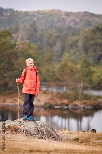 A boy standing on a rock by a lake holding a stick, smiling to camera, Lake District, UK, vertical © Monkey Business