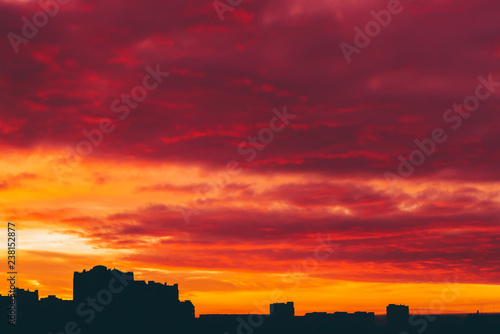 Cityscape with vivid surrealistic dawn. Amazing warm dramatic cloudy sky above dark silhouettes of city buildings. Orange sunlight. Atmospheric background of sunrise in overcast weather. Copy space.