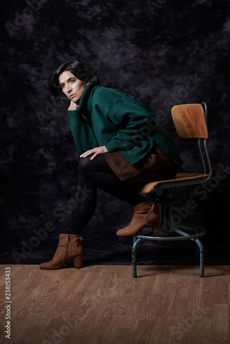 Portrait of a Latin woman in Autumn-Winter outfit sitting on a vintage chair in front of a black textured backdrop.