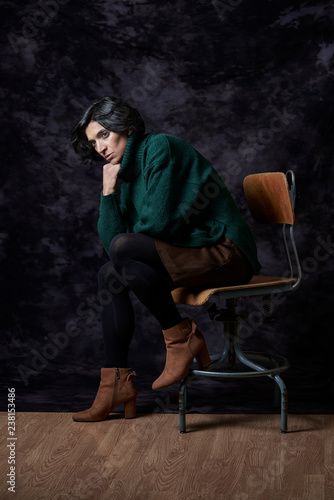 Portrait of a Latin woman in Autumn-Winter outfit sitting on a vintage chair in front of a black textured backdrop.