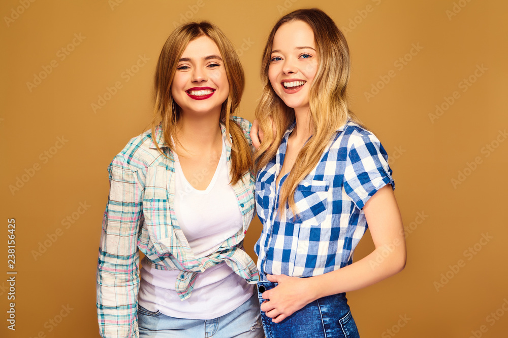Two young beautiful blond smiling hipster girls posing in trendy summer checkered shirt clothes. Carefree women isolated on golden background. Positive models going crazy and hugging Two young beautiful blond smiling hipster girls posing in trendy summer checkered shirt clothes. Carefree women isolated on golden background. Positive models going crazy and hugging