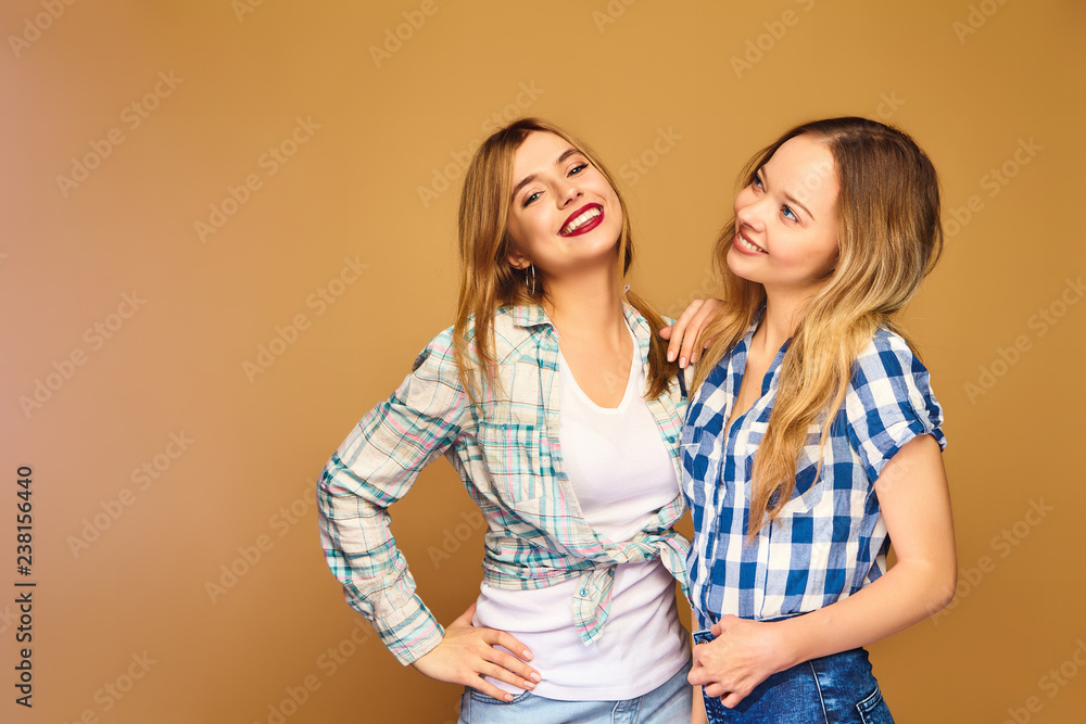 Two young beautiful blond smiling hipster girls posing in trendy summer checkered shirt clothes. Carefree women isolated on golden background. Positive models going crazy and hugging Two young beautiful blond smiling hipster girls posing in trendy summer checkered shirt clothes. Carefree women isolated on golden background. Positive models going crazy and hugging