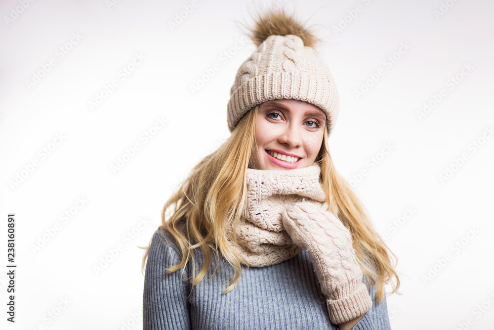 Winter close-up portrait of attractive young blonde woman wearing beige warm knitted hat with fur pompom and scarf snood. Girl looking at camera on white background