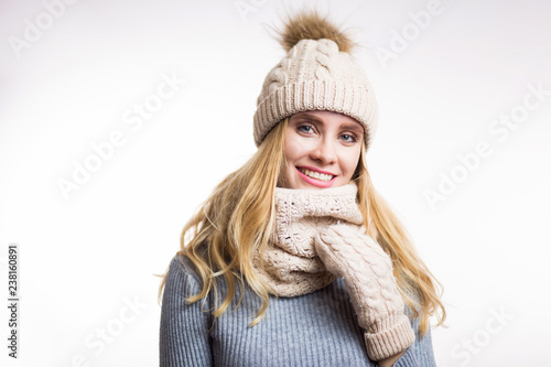 Winter close-up portrait of attractive young blonde woman wearing beige warm knitted hat with fur pompom and scarf snood. Girl looking at camera on white background © liliyabatyrova