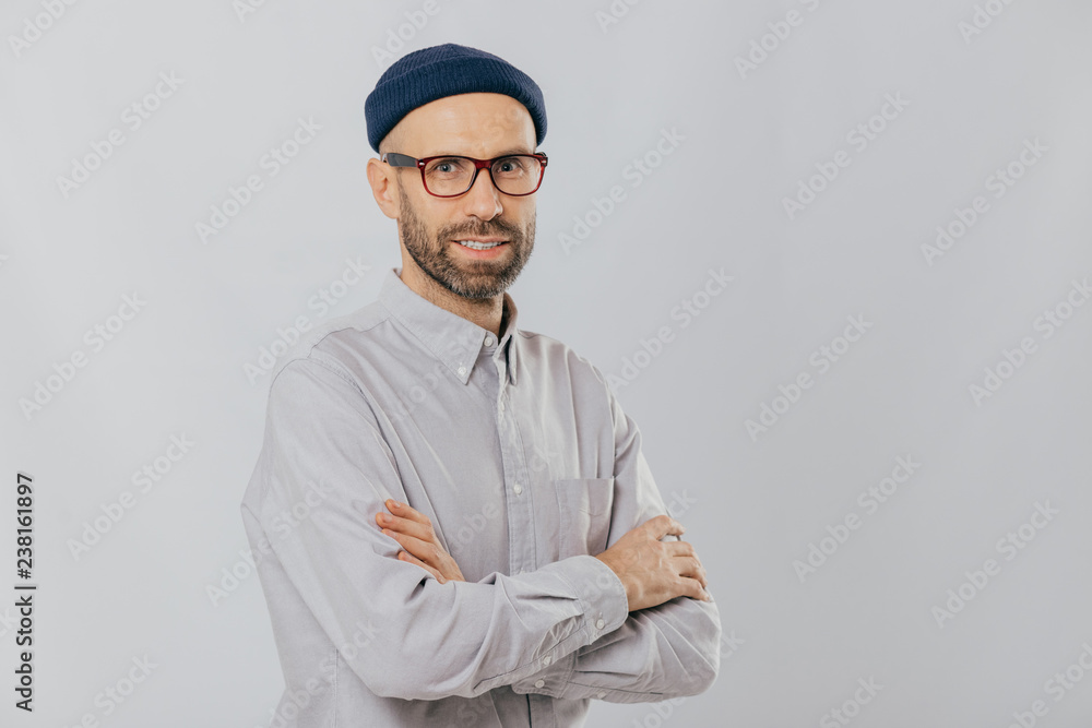 Isolated shot of pleasant looking unshaven male keeps hands crossed, wears stylish hat, optical glasses, models over white background with copy space for your advertising content, enjoys leisure Isolated shot of pleasant looking unshaven male keeps hands crossed, wears stylish hat, optical glasses, models over white background with copy space for your advertising content, enjoys leisure