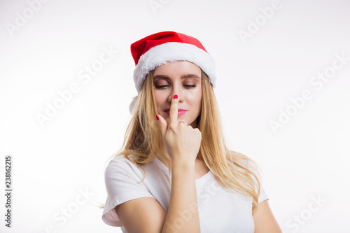 Beautiful funny blonde woman wearing red Santa hat smiling and finger pointing at her nose on white background