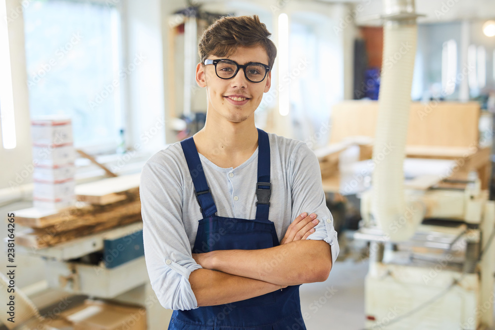 Naklejka premium Smiling confident handsome young joiner in uniform standing in modern workshop and crossing arms on chest while looking at camera
