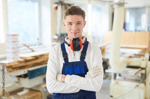 Content confident handsome young carpenter in ear protectors on neck wearing gloves and overall standing in woodworking shop and looking at camera