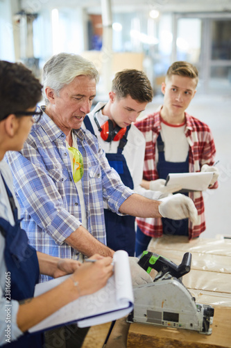 Mature joiner in casual shirt standing at workbench and operating circular saw while demonstrating power tool to young students at practical class