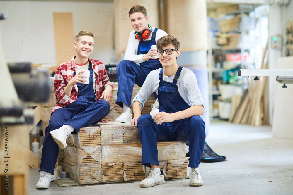 Naklejka premium Wesoło przystojni młodzi stolarze w kombinezonach siedzący na stosie zapakowanych desek parkietowych i patrząc na kamerę, relaksują się podczas przerwy na kawę