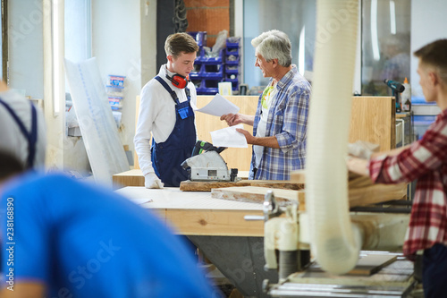 Irritated emotional senior foreman standing at workbench with polish machine and yelling at inexperienced carpenter for mistakes in sketches in factory workshop © pressmaster