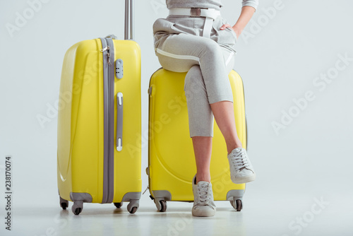cropped view of woman sitting on suitcases with crossed legs on grey background, travel concept