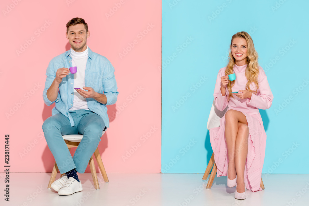 Naklejka premium beautiful smiling couple sitting on chairs, holding toy coffee cups and looking at camera on pink and blue background