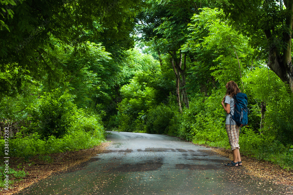 Woman hiker with backpack stands on the road and watches lush green foliage of the tropical trees