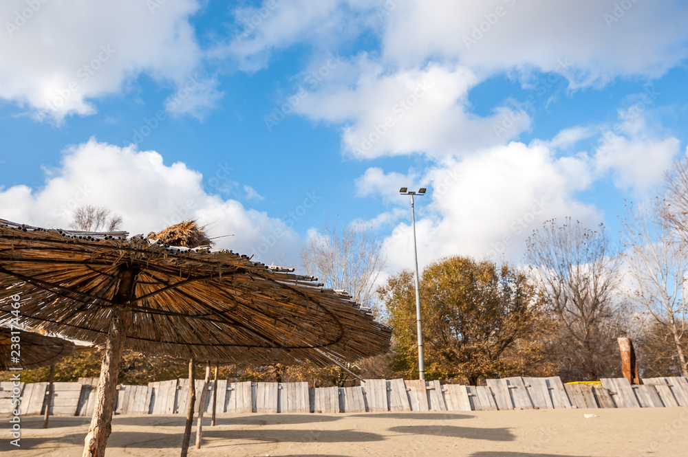 Sunshades or parasols of reed on the empty sandy beach near water with sunny blue sky with clouds for recreational leisure activity and shadow under them Sunshades or parasols of reed on the empty sandy beach near water with sunny blue sky with clouds for recreational leisure activity and shadow under them