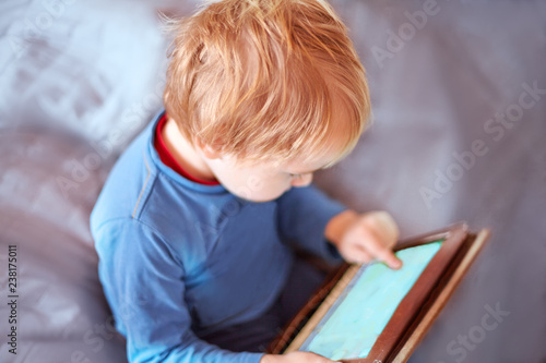 Little caucasian baby boy sits on the sofa using a tablet, touching screen. Red hair, casual wear, indoors, close up, copy space. Children time spending, computerization of youngsters concept.
