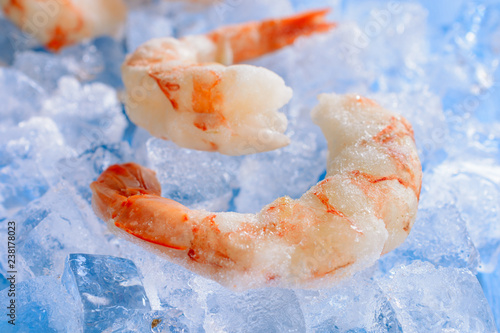 Two boiled frozen shrimps are among the pieces of ice on a blue wooden background