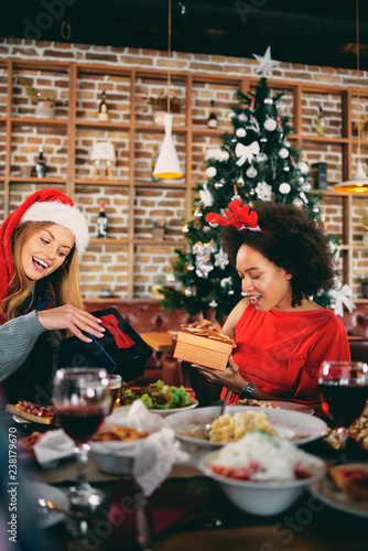 Friends giving gifts to each other while sitting at table. In background Christmas tree. Christmas holidays concept.