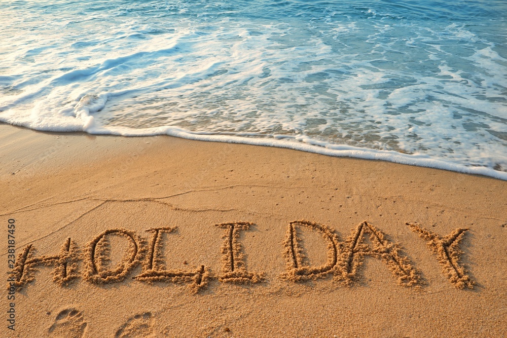 Fototapeta premium Concept HOLIDAY handwriting on sand beach with waves and blue ocean.
