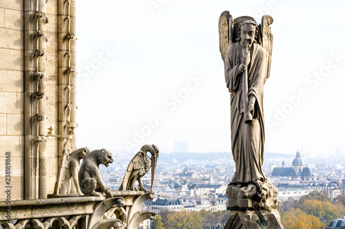 Stone statue of an angel with trumpet on the rooftop of Notre-Dame de Paris cathedral and three chimeras overlooking the city, with church of Saint-Paul-Saint-Louis, vanishing in the mist.