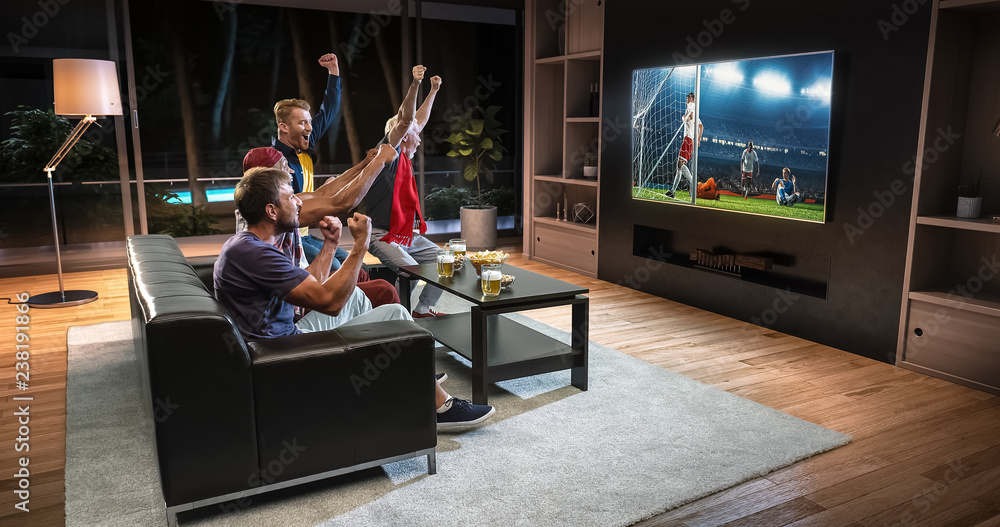 Group of fans are watching a soccer moment on the TV and celebrating a goal, sitting on the couch in the living room.