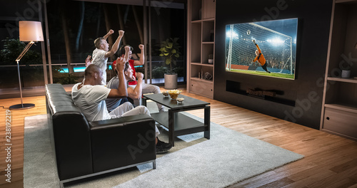 Group of students are watching a soccer moment on the TV and celebrating a goal, sitting on the couch in the living room.