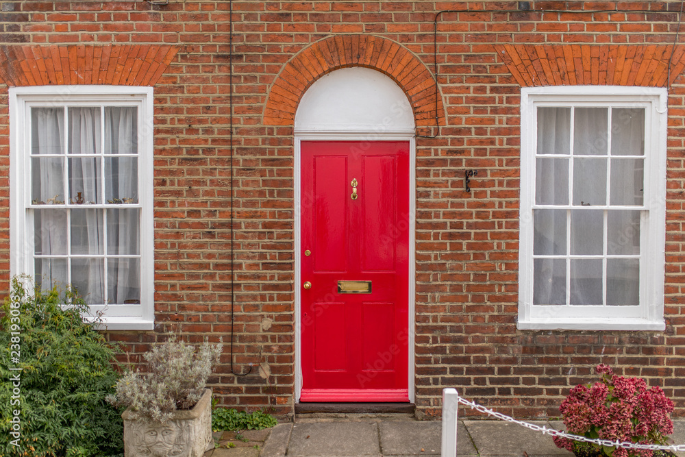 Typical english house facade with red door, two white windows and bricks wall viewed from outdoors.