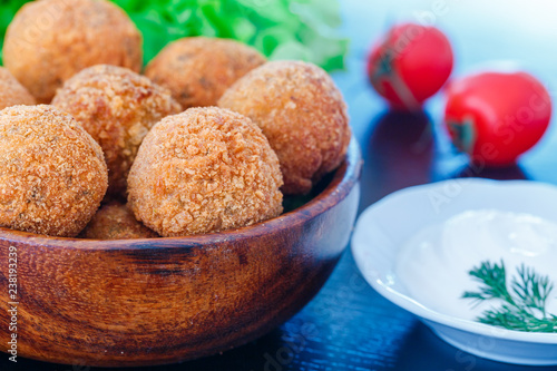 Falafel lies in a wooden plate. On the table lie tomatoes, cucumbers, lettuce, dill, lemon, sour cream.